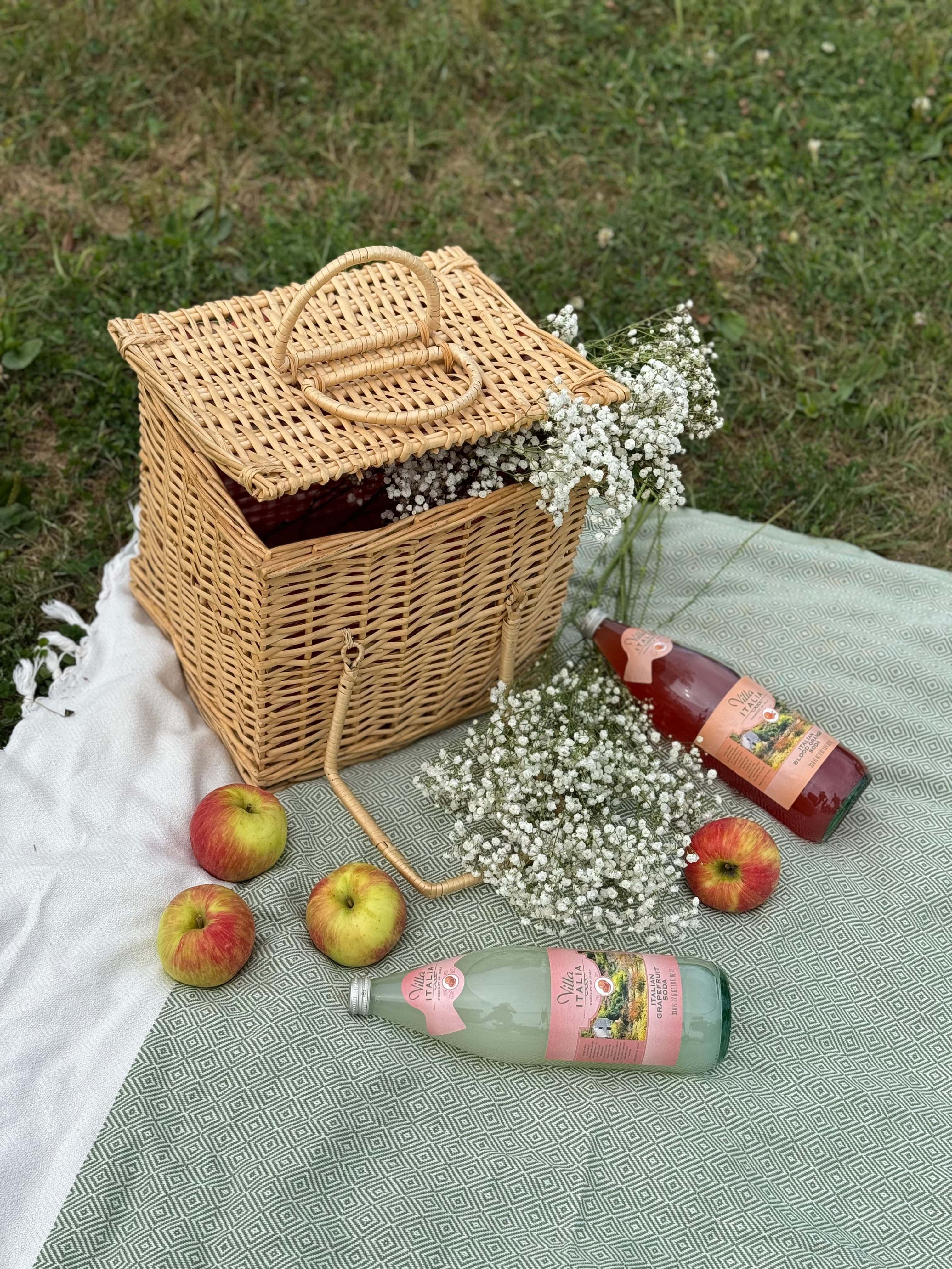Fall picnic with wicker basket, apples, sparkling cider bottles, and baby’s breath flowers on a green blanket.